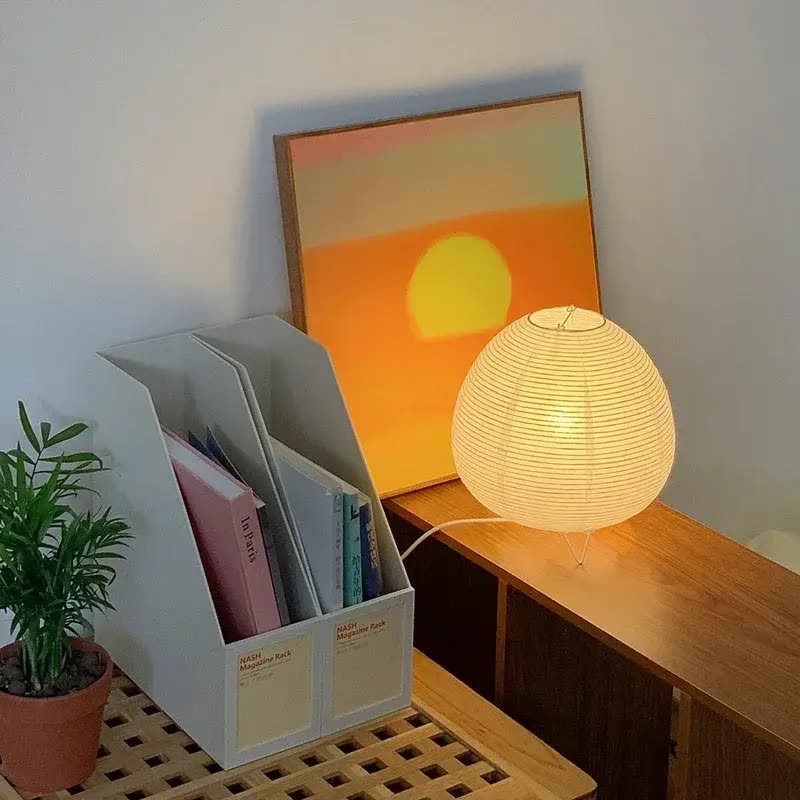 Round cream rice-paper table lamp glowing on a wooden desk beside a plant (seller photo from the AliExpress listing)