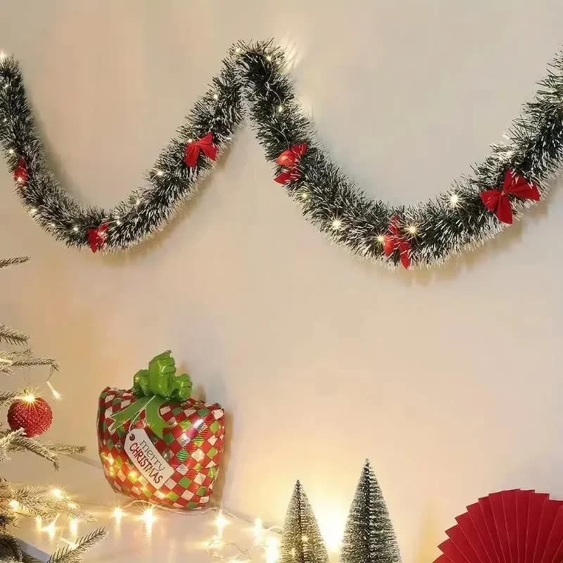 Frosted green tinsel garland with small red bows hung in swags on a white wall above small Christmas trees (seller photo from the AliExpress listing)